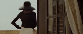 Movie still from “By the Sea” (2015), directed by Angelina Jolie – A woman wearing a black hat standing in front of a window; Medium shot, Low angle
