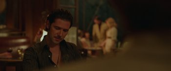 Movie still from “By the Sea” (2015), directed by Angelina Jolie – A man sitting in front of a group of people; Medium shot, Over the shoulder angle