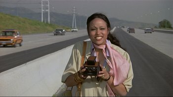 Movie still from “California Suite” (1978), directed by Herbert Ross – A woman holding a camera on the side of a road; Medium shot, High angle