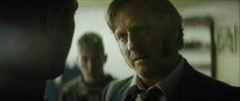 Movie still from “Calm with Horses” (2019), directed by Nick Rowland – A man in a suit and tie looking at the camera with another man behind him; Close Up shot, Over the shoulder angle