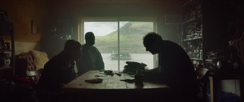 Movie still from “Calm with Horses” (2019), directed by Nick Rowland – Three people sitting at a table in front of a large window; Wide shot, Over the shoulder angle