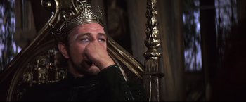 Movie still from “Camelot” (1967), directed by Joshua Logan – A man wearing a gold crown sitting in front of a golden pole; Close Up shot, Low angle