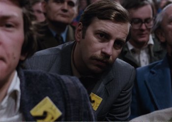 Movie still from “Camera Buff” (1979), directed by Krzysztof Kieslowski – A man in a suit and tie sitting in a crowd of people; Close Up shot, Over the shoulder angle