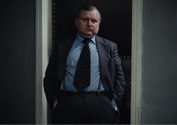 Movie still from “Camera Buff” (1979), directed by Krzysztof Kieslowski – A man in a suit and tie standing in front of a door; Medium shot, Low angle