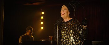 Movie still from “Can You Ever Forgive Me?” (2018), directed by Marielle Heller – A woman in a black and gold outfit is singing; Medium shot, Over the shoulder angle