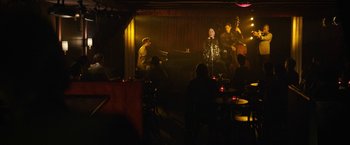 Movie still from “Can You Ever Forgive Me?” (2018), directed by Marielle Heller – A group of people sitting around a room with a microphone; Wide shot, High angle