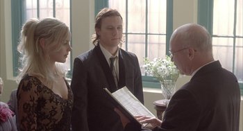 Movie still from “Candy” (2006), directed by Neil Armfield – A man in a suit is holding a book while two women stand next to him; Medium shot, Low angle