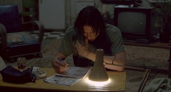 Movie still from “Candy” (2006), directed by Neil Armfield – A person sitting at a table with a lamp on top of a table; Medium shot, High angle