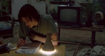 Movie still from “Candy” (2006), directed by Neil Armfield – A person sitting at a table in a room with a lamp; Medium shot, High angle
