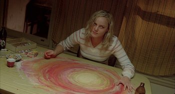 Movie still from “Candy” (2006), directed by Neil Armfield – A woman sitting in front of a table with a painting on top of it; Medium shot, Overhead angle