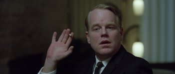 Movie still from “Capote” (2005), directed by Bennett Miller – A man in a black suit and a black tie holding his hand up; Close Up shot, Over the shoulder angle