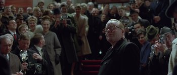 Movie still from “Capote” (2005), directed by Bennett Miller – An older woman is standing in front of a group of people; Close Up shot, Over the shoulder angle