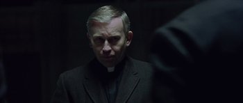 Movie still from “Capote” (2005), directed by Bennett Miller – A man wearing a priest's outfit looking at the camera; Close Up shot, Over the shoulder angle