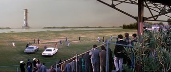 Movie still from “Capricorn One” (1977), directed by Peter Hyams – A group of people standing on top of a grass covered field; Extreme Wide shot, High angle