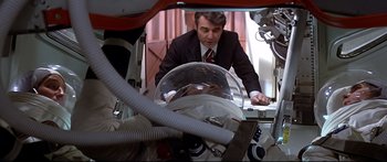 Movie still from “Capricorn One” (1977), directed by Peter Hyams – A man in a suit and tie next to an astronaut helmet; Medium shot, Over the shoulder angle