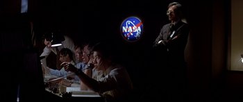 Movie still from “Capricorn One” (1977), directed by Peter Hyams – A group of people sitting at a table in front of a nasa logo; Wide shot, Low angle