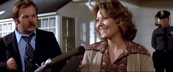 Movie still from “Capricorn One” (1977), directed by Peter Hyams – A woman smiles while standing in front of a microphone; Close Up shot, Over the shoulder angle