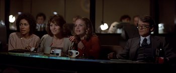 Movie still from “Capricorn One” (1977), directed by Peter Hyams – A group of people sitting at a table with drinks; Medium shot, Over the shoulder angle