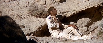 Movie still from “Capricorn One” (1977), directed by Peter Hyams – A man sitting on the ground near some rocks; Medium shot, Overhead angle