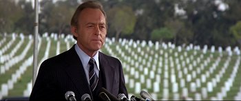 Movie still from “Capricorn One” (1977), directed by Peter Hyams – A man in a suit and tie standing next to microphones in front of a cemetery; Medium shot, Low angle
