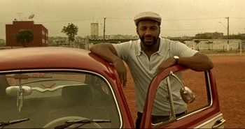 Movie still from “Carandiru” (2003), directed by Hector Babenco – A man standing next to a red car in a field; Medium shot, Low angle
