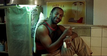 Movie still from “Carandiru” (2003), directed by Hector Babenco – A man sitting in a room holding a remote control; Medium shot, Low angle