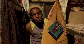 Movie still from “Carandiru” (2003), directed by Hector Babenco – A man holding a surfboard in a room; Medium shot, Low angle