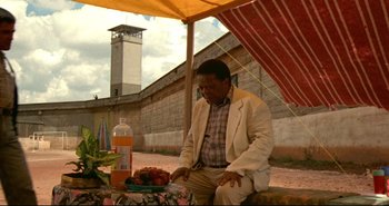 Movie still from “Carandiru” (2003), directed by Hector Babenco – A man sitting under an awning with a bowl of fruit; Wide shot, Over the shoulder angle