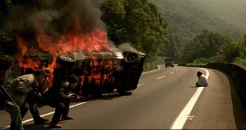 Movie still from “Carandiru” (2003), directed by Hector Babenco – A car that is on fire on the side of the road; Wide shot, High angle