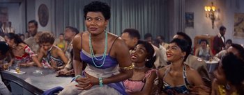 Movie still from “Carmen Jones” (1954), directed by Otto Preminger – A woman sitting next to another woman in front of a crowd; Medium shot, Over the shoulder angle