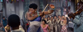 Movie still from “Carmen Jones” (1954), directed by Otto Preminger – An african - american woman in a blue and white dress holding a wooden stick in front of a group of people; Medium shot, High angle