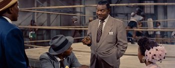 Movie still from “Carmen Jones” (1954), directed by Otto Preminger – A man in a suit and a hat talking to another man in a suit and a hat; Medium shot, High angle