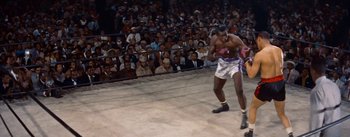 Movie still from “Carmen Jones” (1954), directed by Otto Preminger – A man in white shorts and a pink and white shirt is boxing in front of an audience; Wide shot, High angle