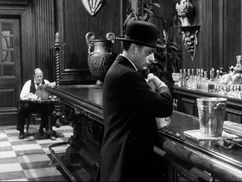 Movie still from “Carrie” (1952), directed by William Wyler – A man standing at a bar in a suit and a hat; Medium shot, Over the shoulder angle