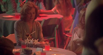 Movie still from “Carrie” (1976), directed by Brian De Palma – A woman sitting at a table with a cake in front of her; Medium shot, Over the shoulder angle