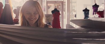 Movie still from “Carrie” (2013), directed by Kimberly Peirce – A woman smiling in front of a dress on display; Medium shot, Over the shoulder angle