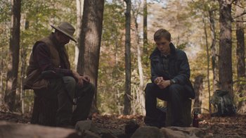Movie still from “The Clovehitch Killer” (2018), directed by Duncan Skiles – Two men sitting in the woods near some rocks; Medium shot, Over the shoulder angle