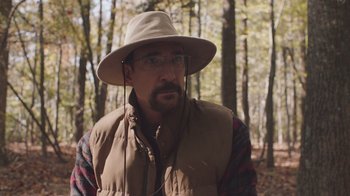 Movie still from “The Clovehitch Killer” (2018), directed by Duncan Skiles – A man with a hat and glasses in the woods; Close Up shot, Over the shoulder angle