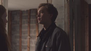 Movie still from “The Clovehitch Killer” (2018), directed by Duncan Skiles – A young man standing in a room next to a brick wall; Close Up shot, Over the shoulder angle