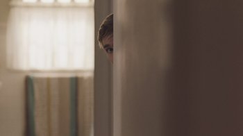 Movie still from “The Clovehitch Killer” (2018), directed by Duncan Skiles – A man peeks out from behind a wall in a bathroom; Medium shot, Over the shoulder angle