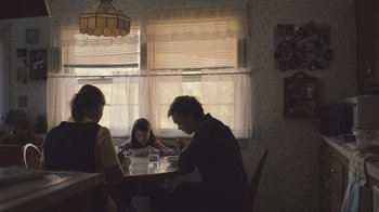 Movie still from “The Clovehitch Killer” (2018), directed by Duncan Skiles – Three people are sitting at a table in a kitchen; Medium shot, Over the shoulder angle