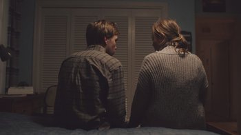 Movie still from “The Clovehitch Killer” (2018), directed by Duncan Skiles – A couple of people sitting on top of a bed next to each other; Medium shot, Over the shoulder angle