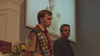 Movie still from “The Clovehitch Killer” (2018), directed by Duncan Skiles – A boy scout standing next to a man in front of a screen; Medium shot, Over the shoulder angle