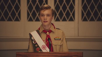 Movie still from “The Clovehitch Killer” (2018), directed by Duncan Skiles – A young boy in a scout uniform stands in front of a wooden table; Medium shot, Over the shoulder angle