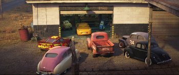 Movie still from “Cars 3” (2017), directed by Brian Fee – A group of old cars parked in front of a garage door; Extreme Wide shot, High angle