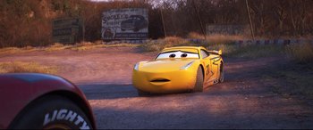 Movie still from “Cars 3” (2017), directed by Brian Fee – A yellow race car is driving down the road; Wide shot, Over the shoulder angle