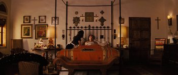 Movie still from “Casa de mi Padre” (2012), directed by Matt Piedmont – A woman sitting on top of a bed next to a dog; Wide shot, High angle