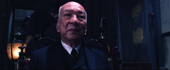 Movie still from “Casshern” (2004), directed by Kazuaki Kiriya – An older man wearing a suit and tie in front of a microphone; Close Up shot, Low angle