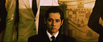 Movie still from “Casshern” (2004), directed by Kazuaki Kiriya – A man in a suit and tie sitting in front of a chair; Close Up shot, Low angle