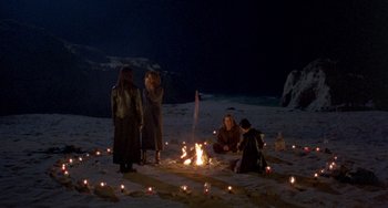 Movie still from “The Craft” (1996), directed by Andrew Fleming – A group of people sitting around a fire on the beach; Wide shot, High angle